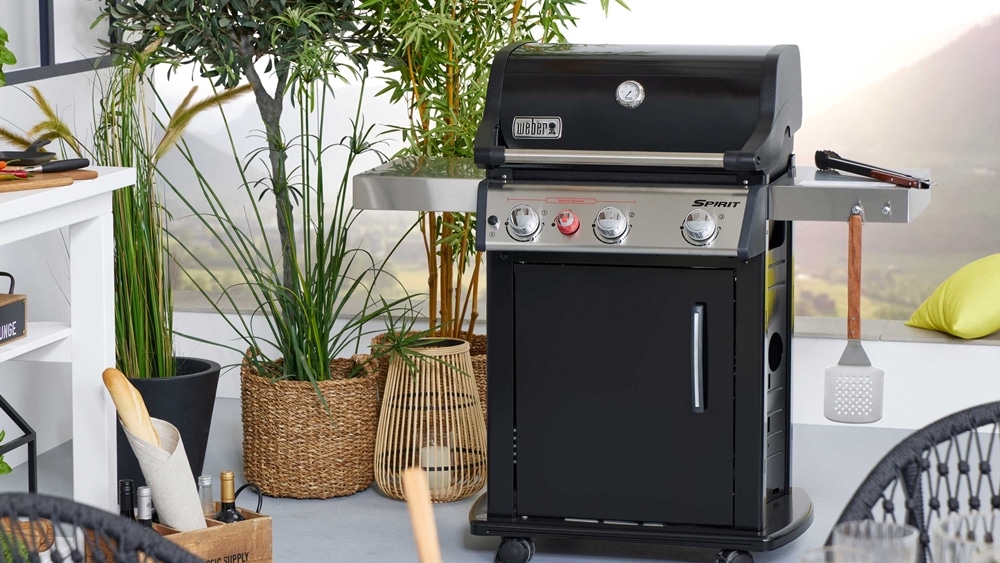 Ein schwarzer Gasgrill mit Utensilien von Möbel Gruber steht auf einer Terrasse, umgeben von Pflanzen und Gartenmöbeln.