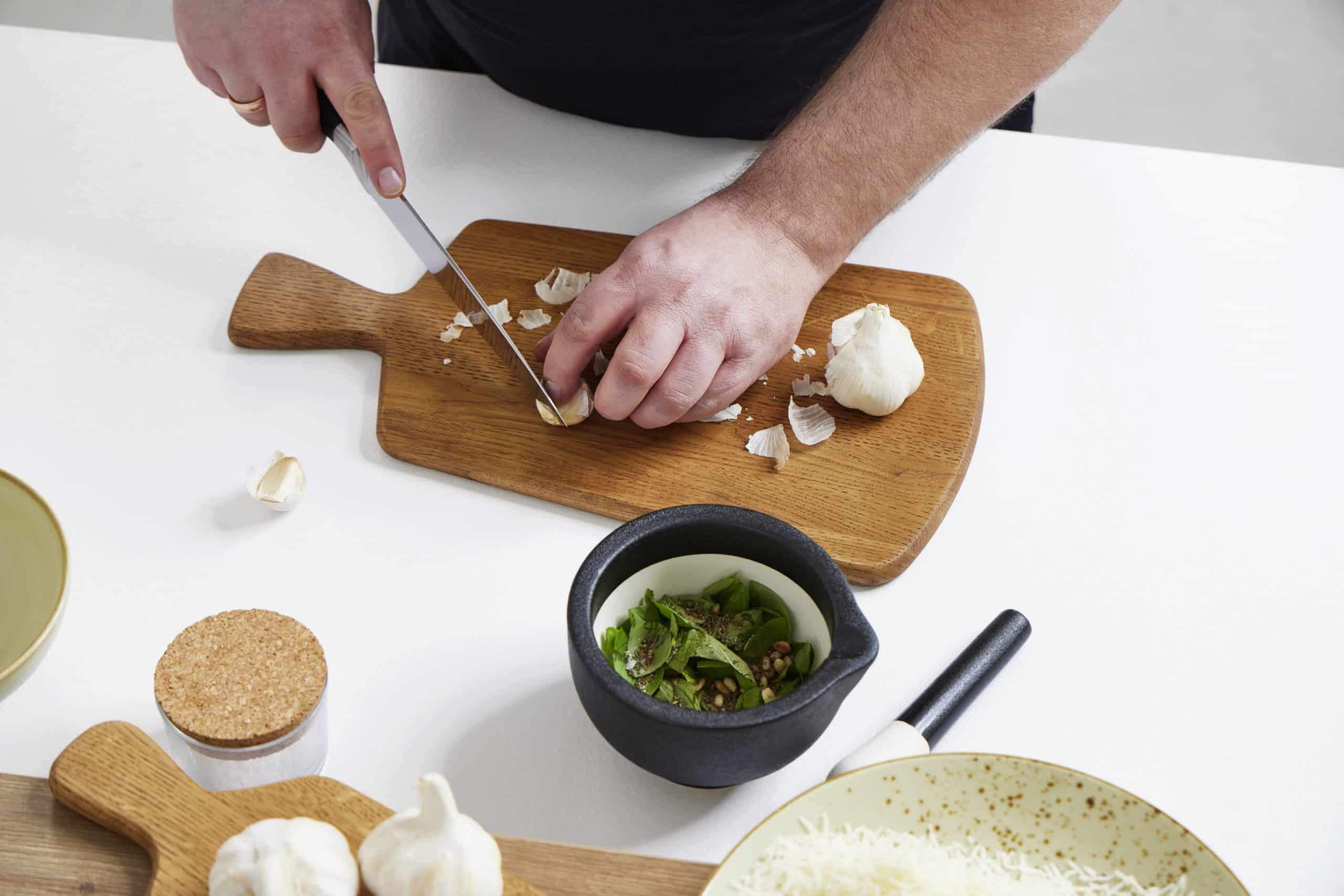 Knoblauch hacken auf einem Brett von Möbel Gruber, umgeben von Schüsseln mit Zutaten und ganzen Zehen.