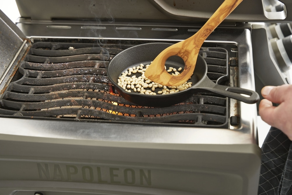 Eine gusseiserne Pfanne mit weißen Bohnen steht auf einem Napoleon-Grill von Möbel Gruber und wird mit einem Holzlöffel umgerührt.