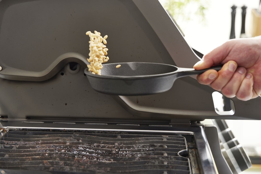 Nahaufnahme einer Hand, Pinienkerne in einer gusseisernen Bratpfanne von Möbel Gruber über einem Außengrill schwenkt.