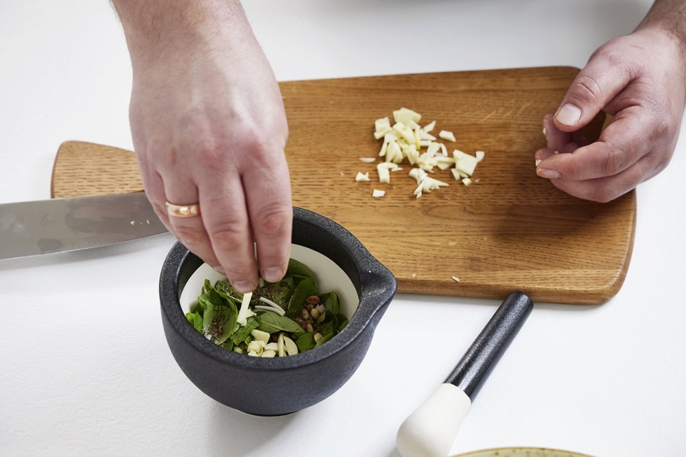 Hände geben gehackten Knoblauch in einen Mörser mit Kräutern, neben einem Holzbrett von Möbel Gruber, auf dem noch mehr Knoblauch und ein Stößel liegen.