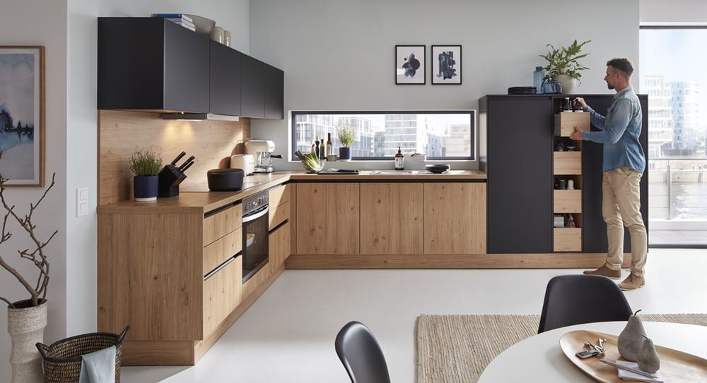 Moderne Küche von Möbel Gruber mit Holzschränken und schwarzen Akzenten. Eine Person neben der Speisekammer genießt große Fenster mit natürlichem Licht.