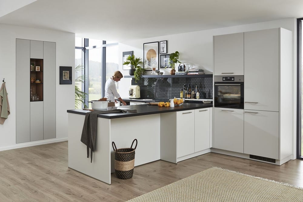 Moderne Küche von Möbel Gruber mit weißen Schränken, schwarzen Arbeitsplatten und einem Korb auf dem Holzboden, während jemand das Essen zubereitet.