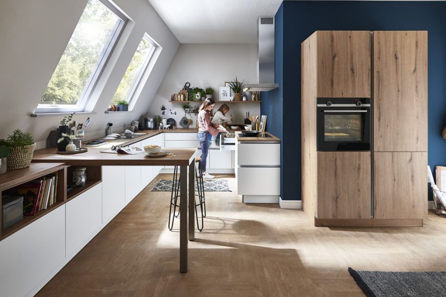 Moderne Küche mit Schränken von Möbel Gruber, einer Frau beim Kochen, großen Fenstern und einer Essinsel mit Hockern.