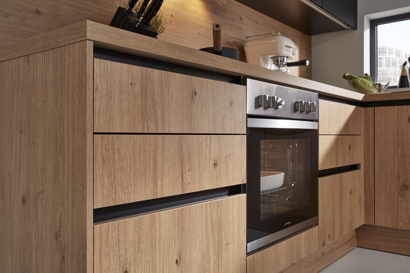 Moderne Küche von Möbel Gruber mit Holzschränken, eingebautem Ofen und einem Küchengerät auf der Arbeitsplatte. Natürliches Licht fällt durch ein kleines Fenster ein.