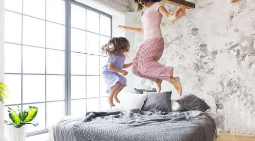 Eine Frau und ein Kind hüpfen fröhlich auf einem Bett von Möbel Gruber in einem hellen, modernen Schlafzimmer mit großen Fenstern und einer strukturierten Wand.