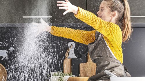 Ein Kind im gelben Pullover wirft fröhlich Mehl in eine Küche von Möbel Gruber, im Hintergrund sind eine Schüssel und Schneidebretter zu sehen.