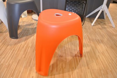 Orangefarbener Kunststoffhocker mit herzförmigem Ausschnitt auf der Oberseite, auf einem Holzboden in einer Umgebung von Möbel Gruber platziert.