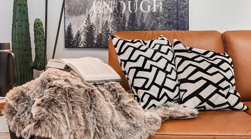 Gemütliches Wohnzimmer von Möbel Gruber mit Ledersofa, geometrischen Kissen, Kunstpelzüberwurf, Stehlampen, Kakteen und Bergkunst.