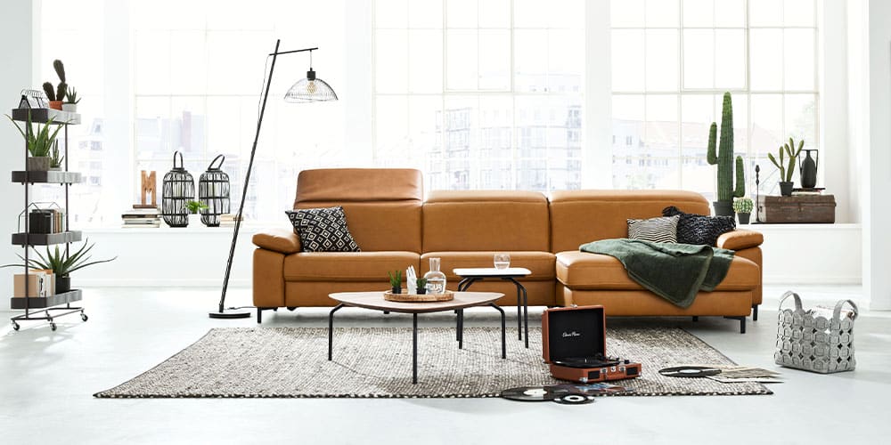 Helles Wohnzimmer mit hellbraunem Sofa, Stehlampe, Tischen und Pflanzen. Große Fenster im Hintergrund.