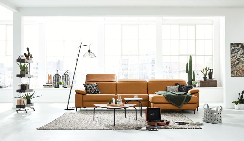 Helles Wohnzimmer mit hellbraunem Sofa, Stehlampe, Tischen und Pflanzen. Große Fenster im Hintergrund lassen natürliches Licht herein.