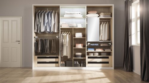 Ein aufgeräumter, offener Kleiderschrank von Möbel Gruber präsentiert hängende Kleidung, gefaltete Hemden, Hosen und Schuhe in einem hellen Raum.
