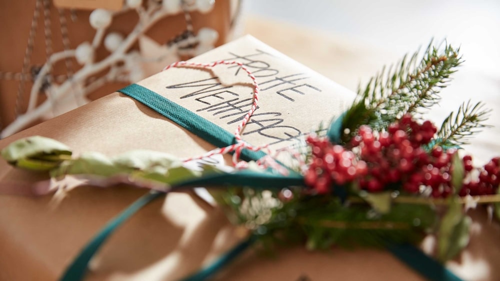 Ein in braunes Papier eingewickeltes Geschenk mit der grünen Schleife von Möbel Gruber, geschmückt mit festlichen roten Beeren und immergrünen Zweigen.
