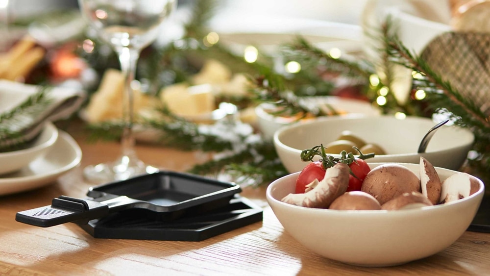 Ein festlicher Tisch von Möbel Gruber mit Schalen voller Pilze, Kirschtomaten und anderen Gerichten, geschmückt mit immergrünen Zweigen und Lichtern.