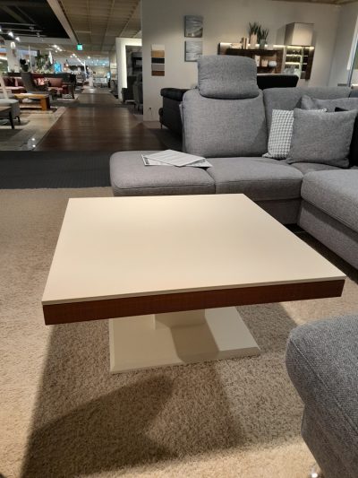 Moderner Ausstellungsraum mit einem beigen Couchtisch von Möbel Gruber und einem grauen Sofagarnitur auf Teppichboden inmitten anderer Ausstellungsstücke.