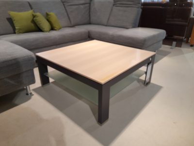 Ein moderner Couchtisch von Möbel Gruber mit Glasablage, vor einem grauen Ecksofa mit grünen Kissen.