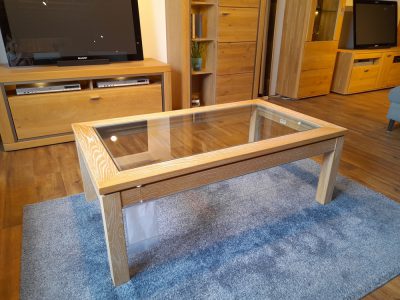 Ein Couchtisch mit Glasplatte von Möbel Gruber steht auf einem blauen Teppich in einem Wohnzimmer mit Holzmöbeln und Fernseher.