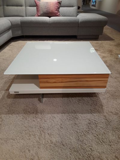 Der Couchtisch mit Glasplatte und Holzseiten steht auf einem beigen Teppich in einem Wohnzimmer mit einem grauen Sofa.