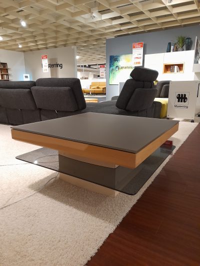 Moderne Wohnzimmerausstellung mit Musterring-Couchtisch, grauen Sofas und abstrakter Kunst, präsentiert bei Möbel Gruber.