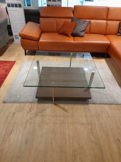 Ein moderner Couchtisch aus Glas von Ronald Schmitt auf einem hellen Teppich ergänzt das orangefarbene Ledersofa von Möbel Gruber.