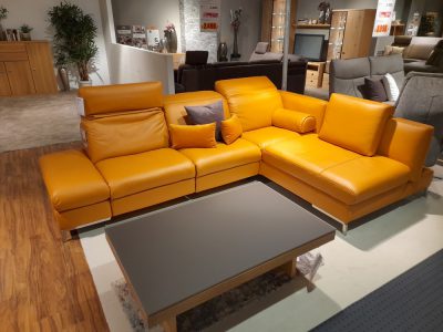 Orangefarbenes Ledersofa „Himolla Polstergarnitur“ mit Kissen neben einem modernen Couchtisch in einem stilvollen Ausstellungsraum.
