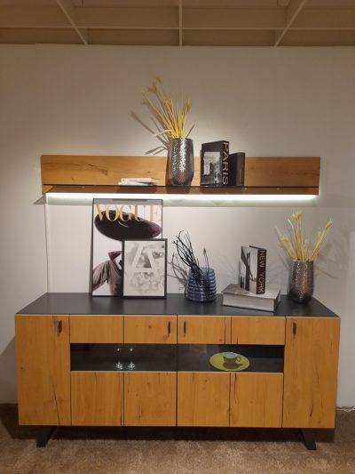 Interliving-Sideboard mit Büchern, Vasen, Blumen und einer Zeitschrift auf einem modernen Regal mit sanfter Beleuchtung darüber.