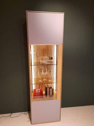 Ein Interliving-Standelementschrank enthält Bücher, Weingläser und eine Flasche auf beleuchteten Glasregalen vor einer dunklen Wand.