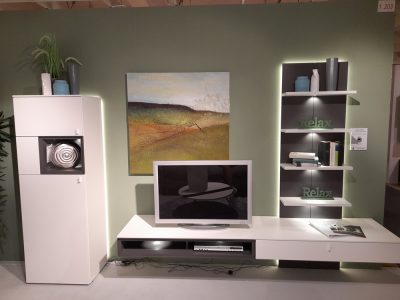 Wohnwand von Modern Möbel Gruber verfügt über einen wandmontierten Fernseher, Regale, Kunstwerke, Dekorationsgegenstände und einen hohen Schrank an einer grünen Wand.