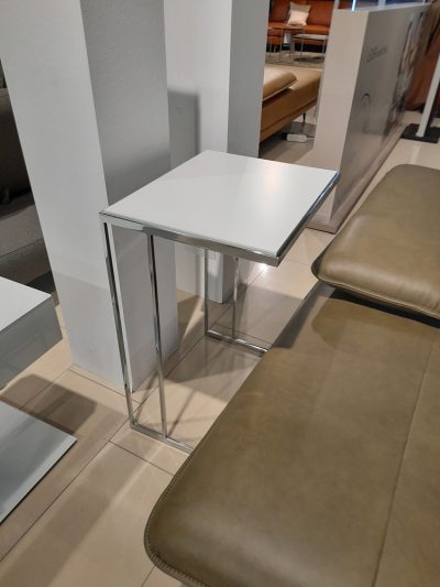 Der Beistelltisch Gallery M, ein kleiner weißer Tisch mit Chromgestell, steht neben einer Lederbank im Showroom von Möbel Gruber.