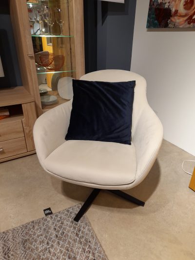 Ein beiger Sessel mit dunkelblauem Kissen steht neben einem Holzschrank mit Glasböden von Möbel Gruber.