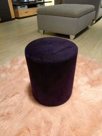 Ein runder Hocker aus lila Samt steht auf einem flauschigen rosa Teppich in einem Wohnzimmer mit einem grauen Sofa im Hintergrund.