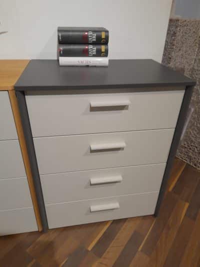 Eine schlichte Kommode mit vier Schubladen, einer dunklen Tischplatte mit gestapelten Büchern, auf einem Holzboden.