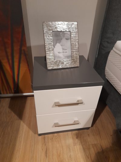 Eine Nachtkonsole mit zwei Schubladen und einer grauen Platte hält ein silbergerahmtes Foto auf dem Holzboden neben dem Bett.
