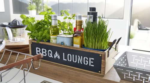 Eine Holzkiste von Möbel Gruber mit der Aufschrift „BBQ Lounge“ enthält Kräuter, Olivenöle und Gewürze auf einer Küchentheke.