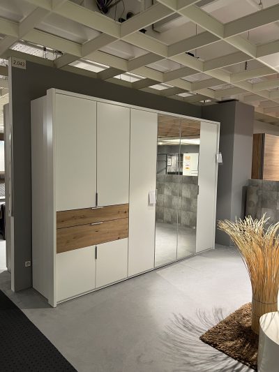 Schlafzimmerschrank mit Spiegeltüren und Holzschubladen in einem Ausstellungsraum, vor einer grauen Wand mit Deckenpaneelen.