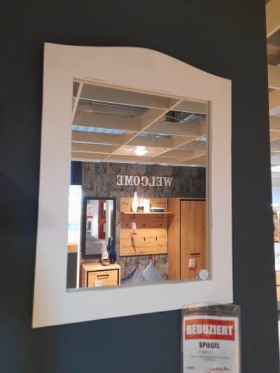 Der Spiegel spiegelt den Ausstellungsraum von Möbel Gruber wider, mit einer Holzvitrine und einem „WELCOME“-Schild an der dunklen Wand.