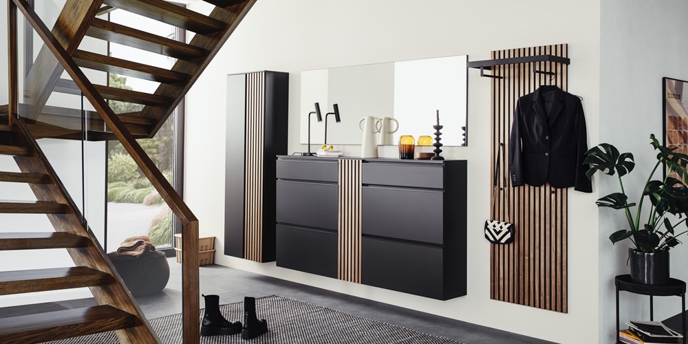 Der moderne Eingangsbereich von Möbel Gruber verfügt über schwarze Schränke, einen großen Spiegel, Holzakzente und eine Treppe mit Garderobe und Lampendekor.