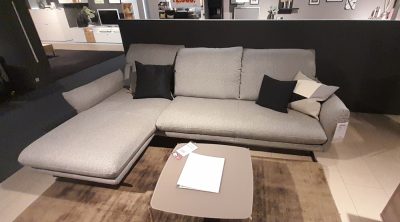 Im Showroom von Möbel Gruber ist die W.Schillig Polstergarnitur mit einem quadratischen Couchtisch auf einem braunen Teppich zu sehen.