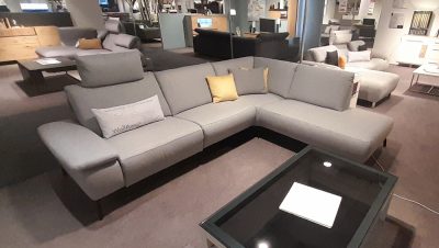 Graues L-Sofagarnitur von Möbel Gruber, mit gelbem Kissen, neben einem schwarzen Couchtisch aus Glas.