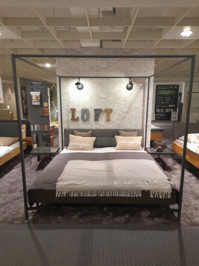 Ein modernes Schlafzimmer von Möbel Gruber verfügt über ein Himmelbett, dekorative Kissen und die Buchstaben „LOFT“ vor einer Backsteinwand im Hintergrund.