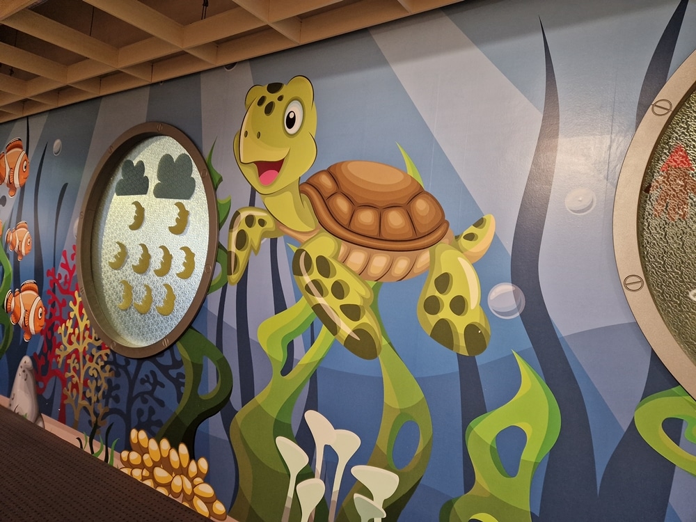 Farbenfrohes Wandbild einer Zeichentrick-Schildkröte, die mit Fischen und Meerespflanzen unter Wasser schwimmt, betrachtet durch ein rundes Fenster.