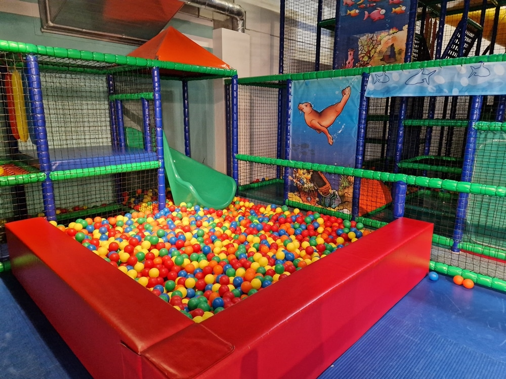 Farbenfroher Indoor-Spielplatz mit Bällebad, Rutsche und von Netzen umgebenen Klettergerüsten in einem gepolsterten Bereich.