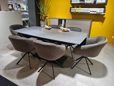 Modernes Esszimmer mit grauem Tisch, Stühlen von Möbel Gruber und gelber Wand. Zur Einrichtung gehören Pflanzen und Geschirr.
