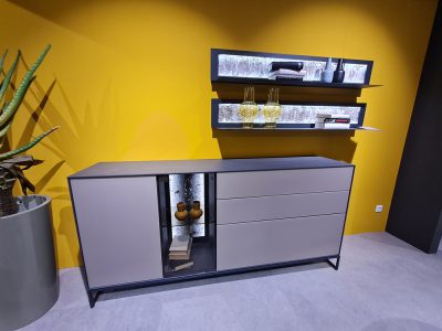 Ein modernes Sideboard von Hartmann mit Dekor oben vor einer gelben Wand, darüber befinden sich Regale mit Glaswaren.
