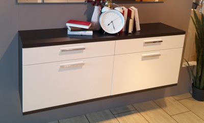 Der an der Wand montierte Schuhschrank mit vier Schubladen, einer Uhr und Büchern darauf steht neben einer Pflanze auf gefliestem Boden.