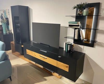 Ein gut beleuchteter Innenraum mit dem modernen Wohnzimmerset „Wohnwand“ von Möbel Gruber mit Fernseher, Regalen und Büchern.