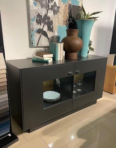 In einem modernen Showroom von Möbel Gruber wird ein schwarzes Sideboard mit Vasen, Büchern und Geschirr gezeigt.