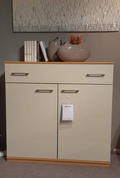 Eine beige Kommode von Möbel Gruber mit Büchern und Vasen, vor einer grauen Wand mit strukturiertem Kunstwerk darüber.