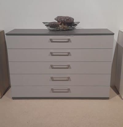 Eine moderne, graue Kommode mit fünf Schubladen, eleganten Griffen und einem dekorativen Korb vor einer weißen Wand.