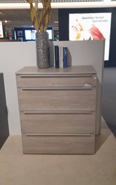 Die Kommode von Möbel Gruber mit vier Schubladen wird mit einer silbernen Vase und Büchern in einer Showroom-Umgebung geschmückt.